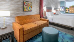 A hotel room with a sofa, a bed, a round ottoman, a nightstand, a lamp, and a painting on the wall.