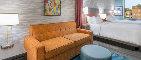 A hotel room with a sofa, a bed, a round ottoman, a nightstand, a lamp, and a painting on the wall.