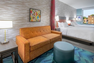 Une chambre d’hôtel avec un canapé, un lit, un pouf rond, une table de chevet, une lampe et un tableau au mur.