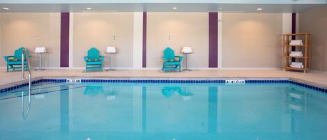 A swimming pool with a clear blue water surface, surrounded by turquoise chairs and a wooden shelf with towels.