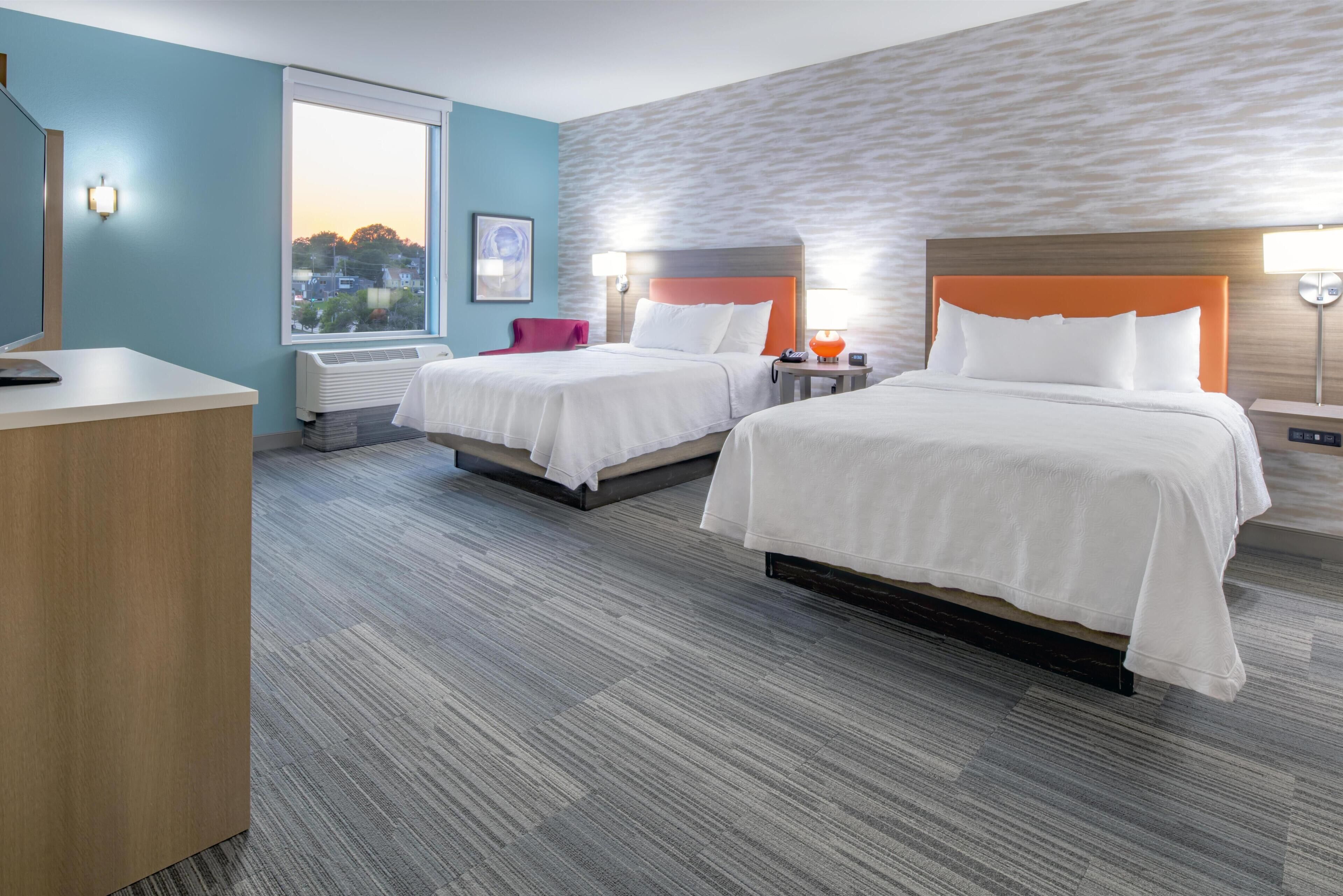 A hotel room with two beds, a desk, a chair, a window with a view, and a wall with a textured pattern.