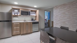 A modern kitchen with a refrigerator, microwave, and dishwasher, a dining table with chairs, and a view of a blue room through an open door.