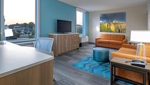 A modern hotel room with a large window, a flat-screen TV, a sofa, a desk, and a painting on the wall.