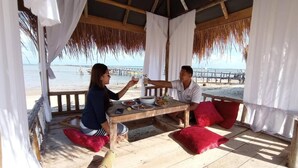 A wooden deck with a table and chairs, overlooking a beach with a pier in the background. - Hello Bintan Beach Cottages (Bintan)