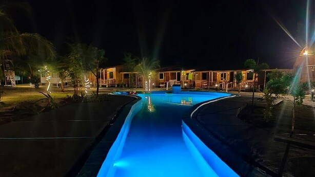 A nighttime view of a resort with a central pool illuminated by blue lights, surrounded by palm trees and a building with outdoor seating. - Hello Bintan Beach Cottages (Bintan)
