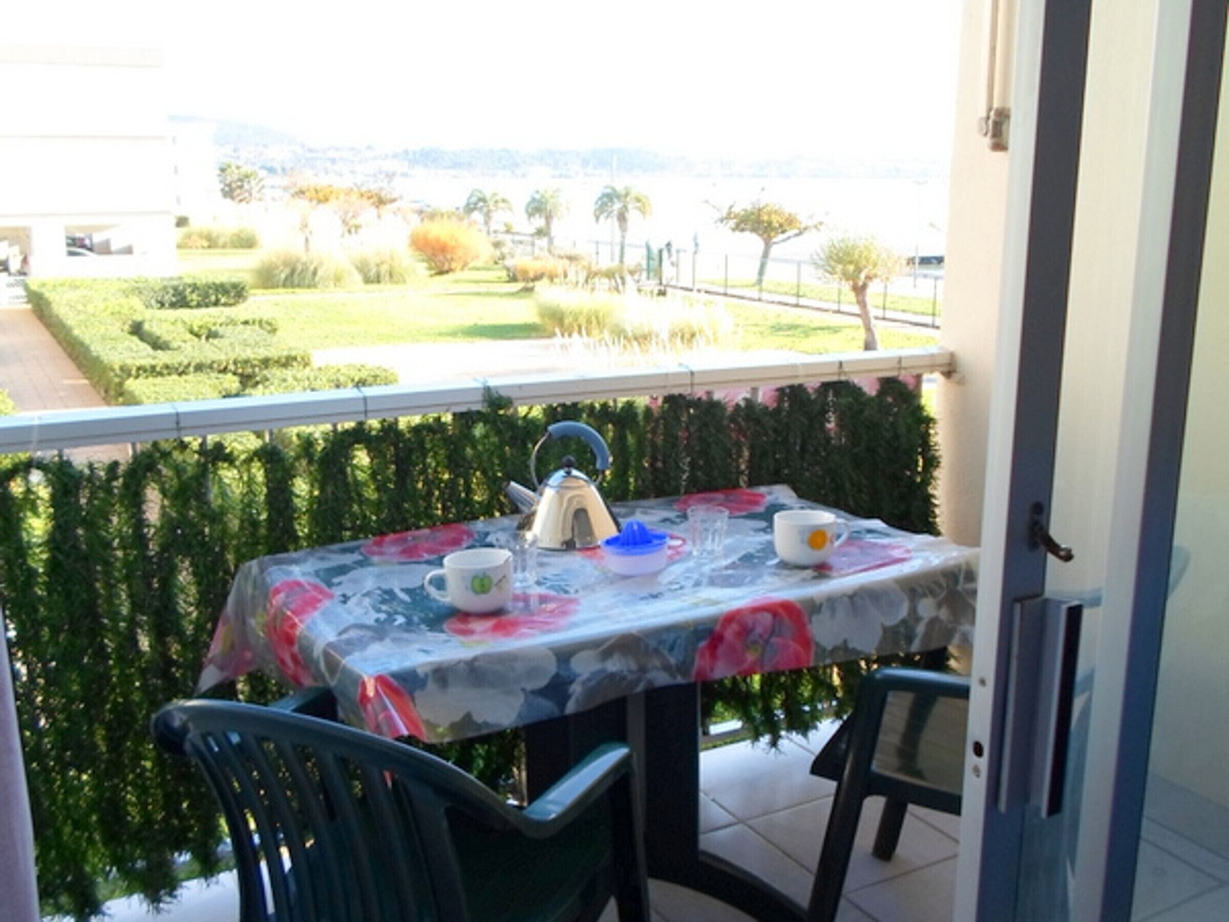 Un balcón con una mesa lista para dos personas, con mantel floreado, dos sillas y una tetera.