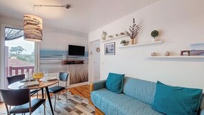 Een gezellige woonkamer met een bank, een eettafel en een aan de muur bevestigde televisie. Er zijn planken met decoratieve objecten en uitzicht op de omgeving.
