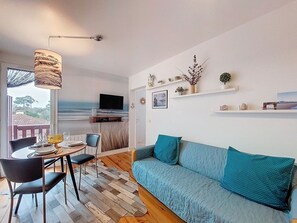 A cozy living room with a sofa, a dining table, and a TV mounted on the wall. There are shelves with decorative items and a view of the outdoors.