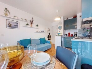 Un apartamento moderno con sala de estar que incluye un sofá azul, una mesa de comedor con un tazón de vidrio y copas de vino, y una cocina con gabinetes azules y encimera blanca.