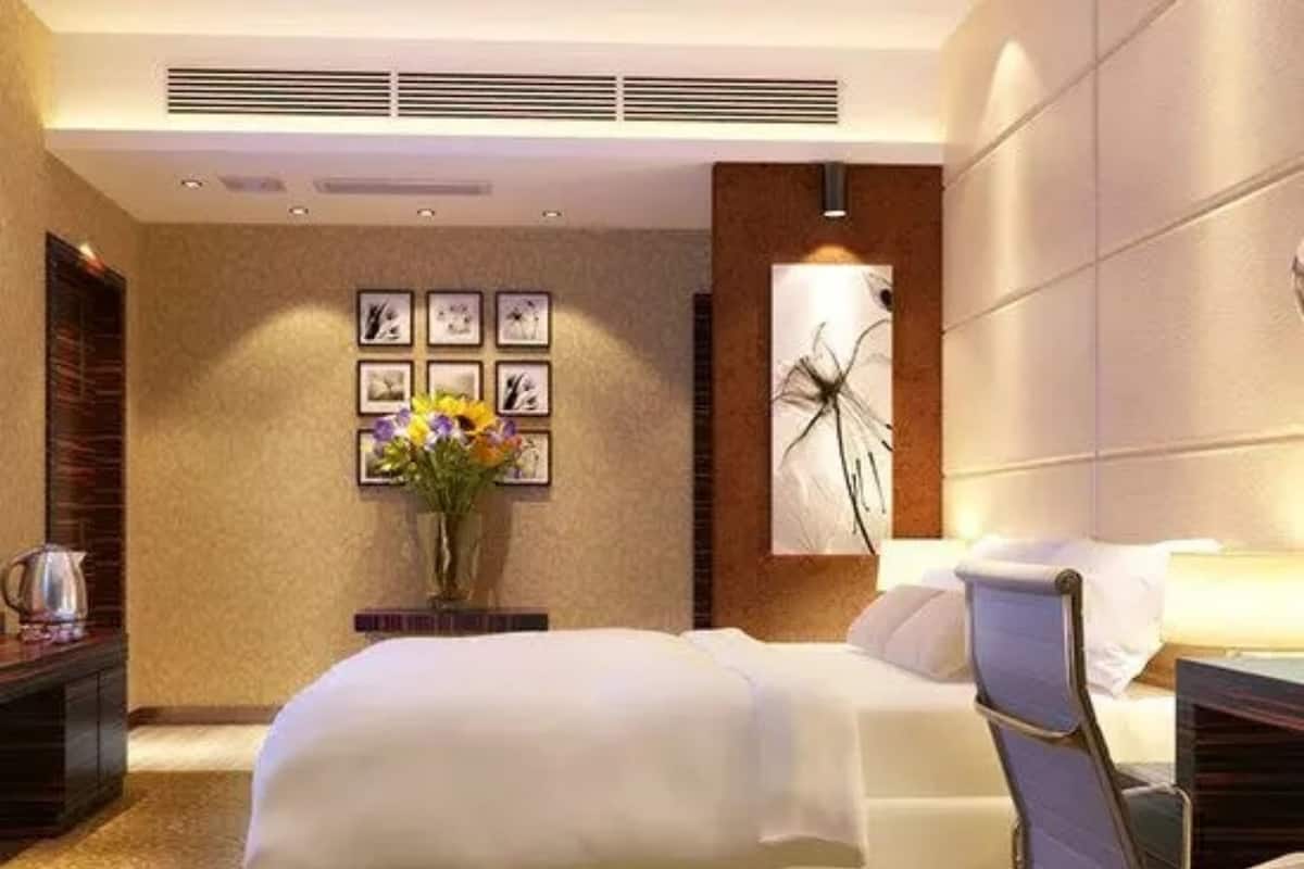 Habitación de hotel con una cama grande, una mesita de noche, un florero con flores, un cuadro colgado en la pared y una rejilla en el techo.
