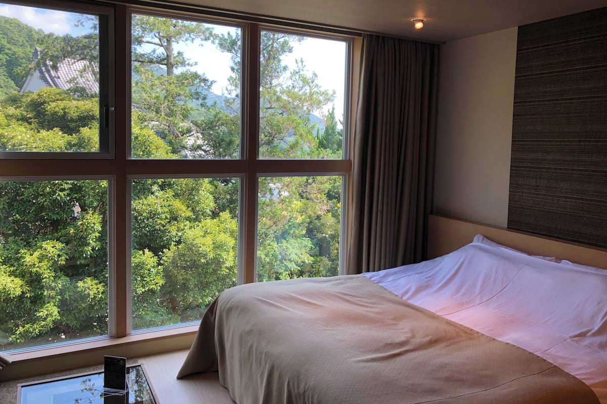 A bedroom with a large window, a bed, and a view of greenery.