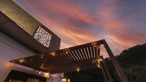 Un edificio moderno con una pérgola de madera y luces colgantes, al atardecer.