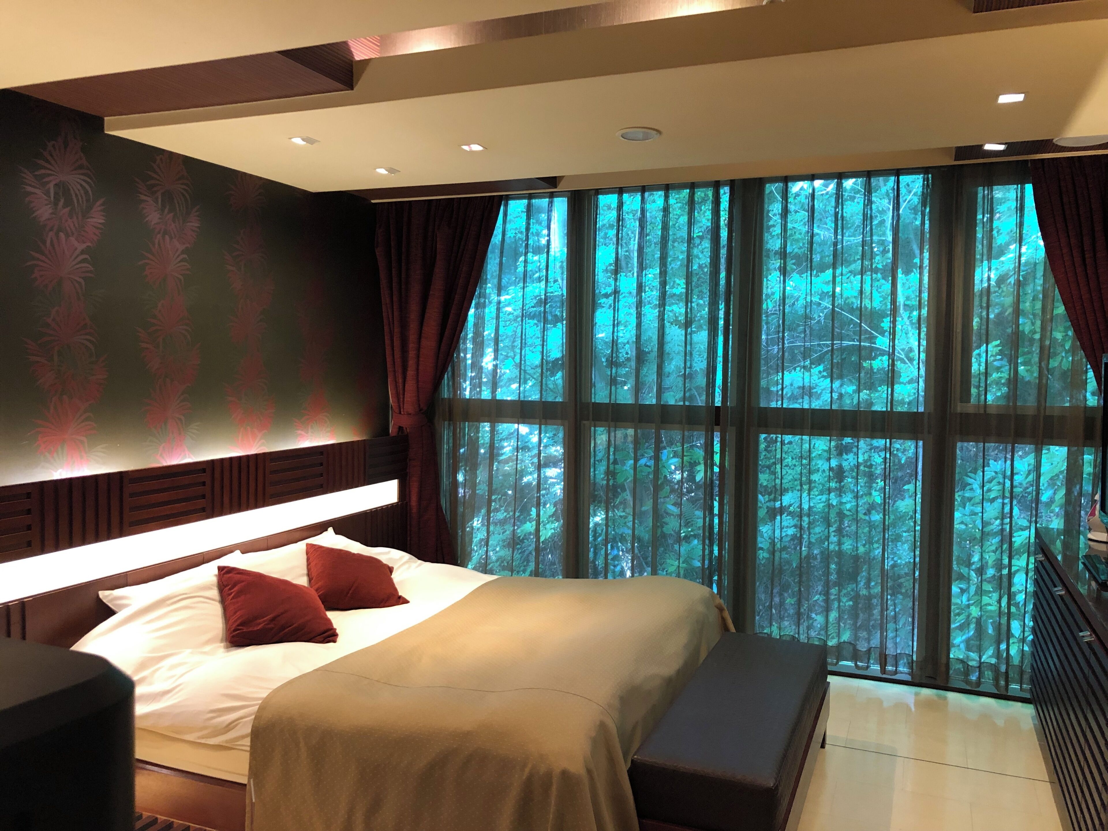 A bedroom with a large bed, a bench, a nightstand, and a view of a forest through large windows.
