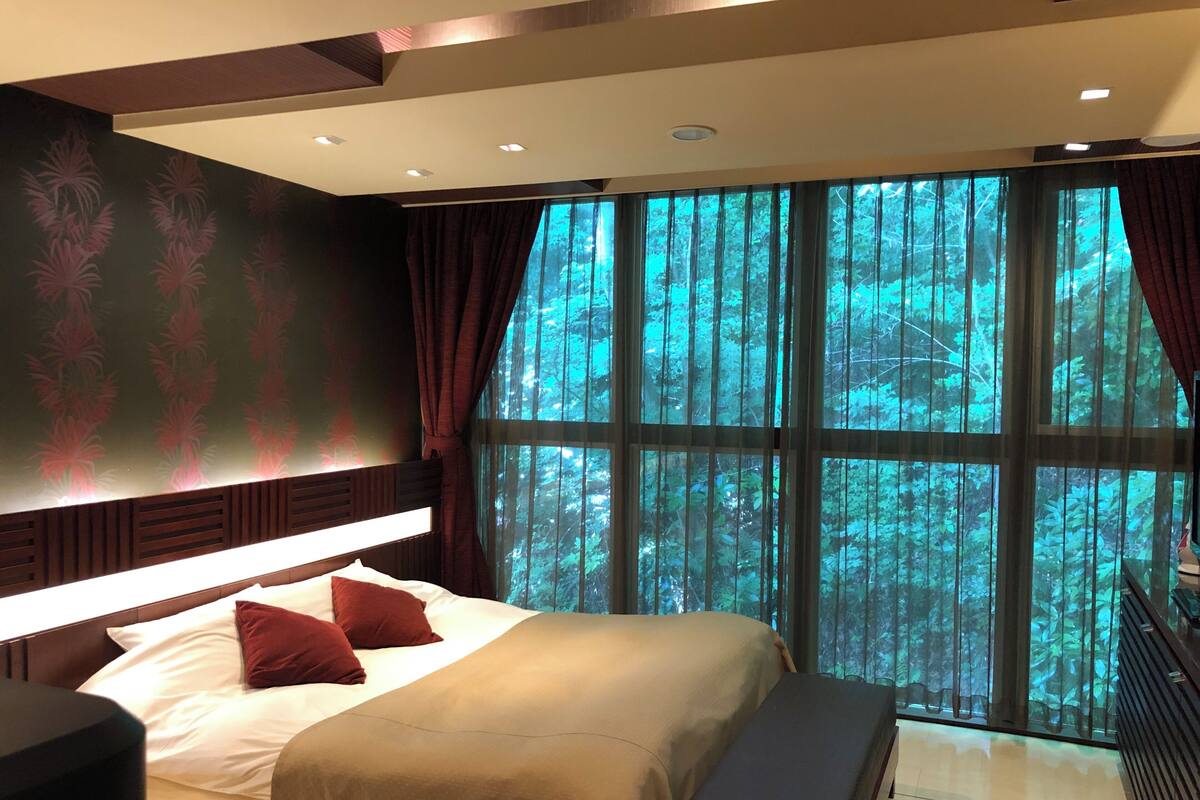 A bedroom with a large bed, a bench, a nightstand, and a view of a forest through large windows.