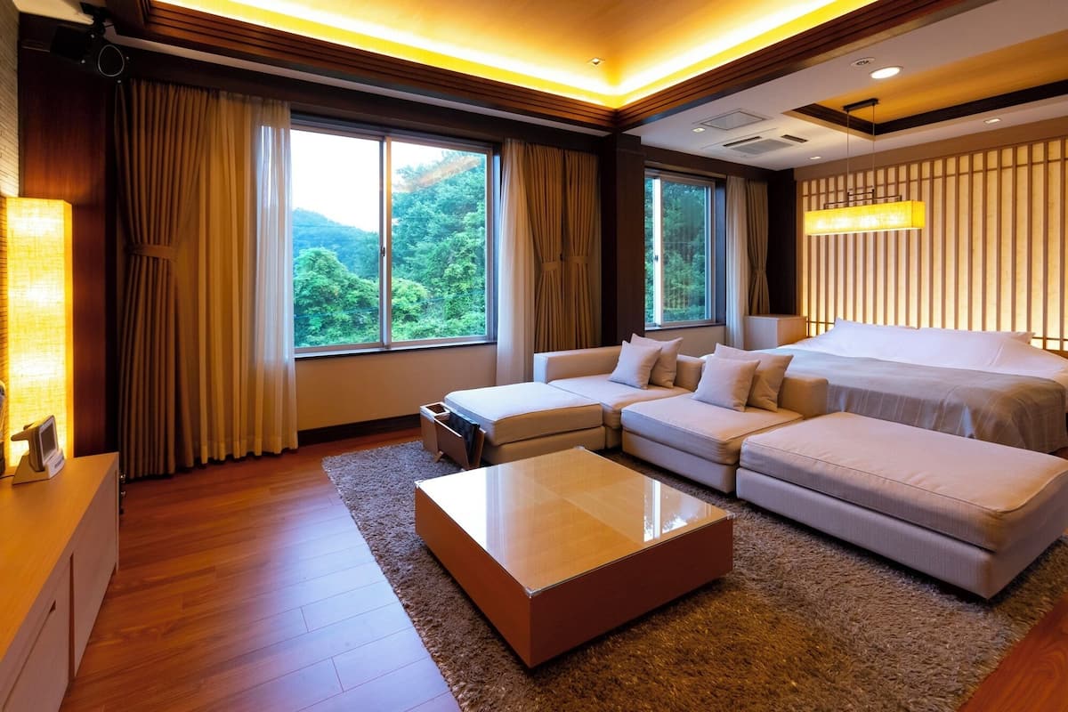 A modern hotel room with a large bed, two sofas, a coffee table, and large windows offering a view of the outdoors.
