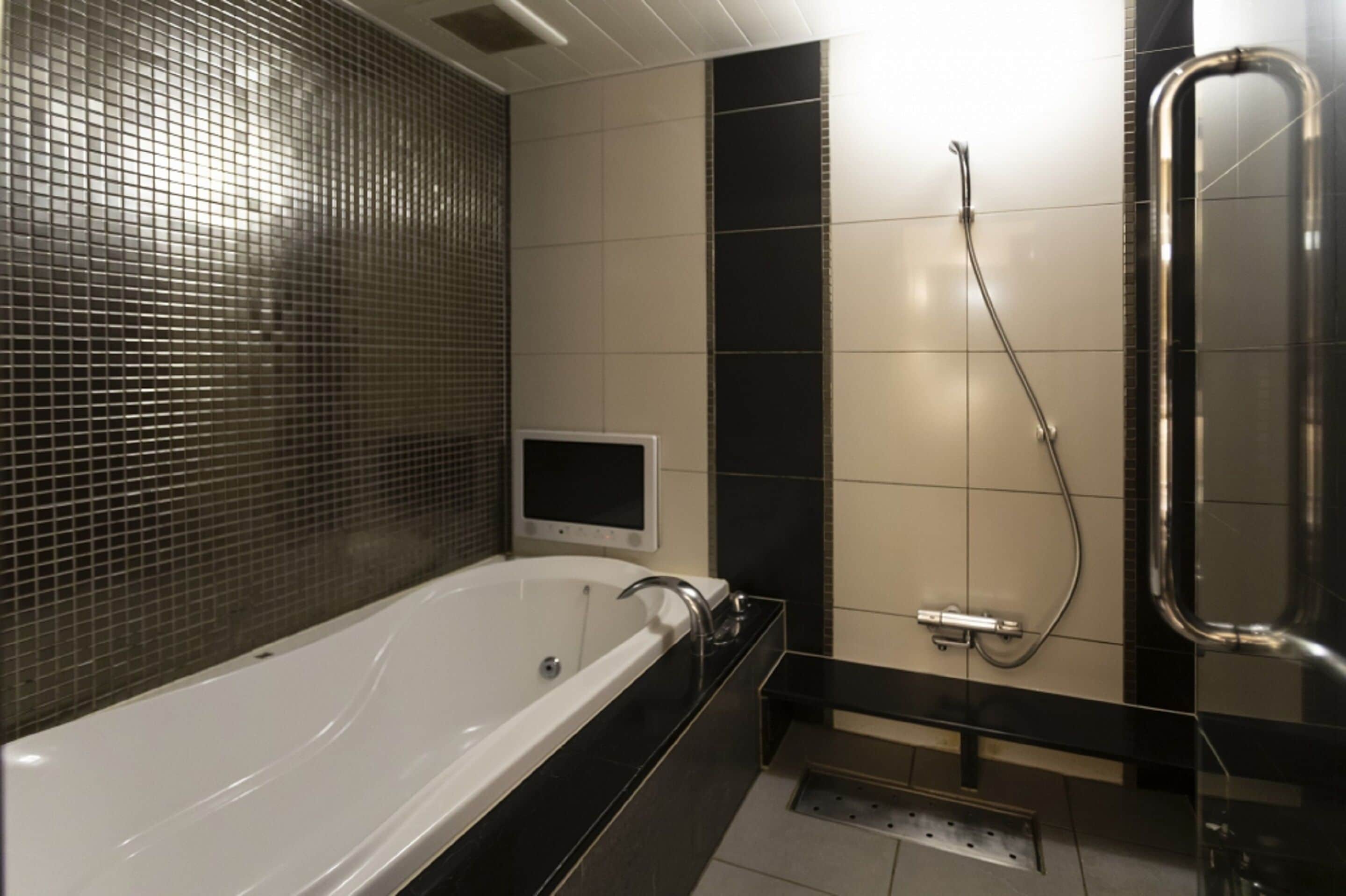 A modern bathroom with a large bathtub, a handheld shower, and a small screen mounted on the wall.