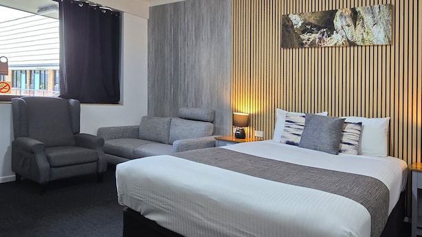 A hotel room with a large bed, two armchairs, a nightstand with a lamp, and a wooden panel wall. - Archer Park Motel (Rockhampton)