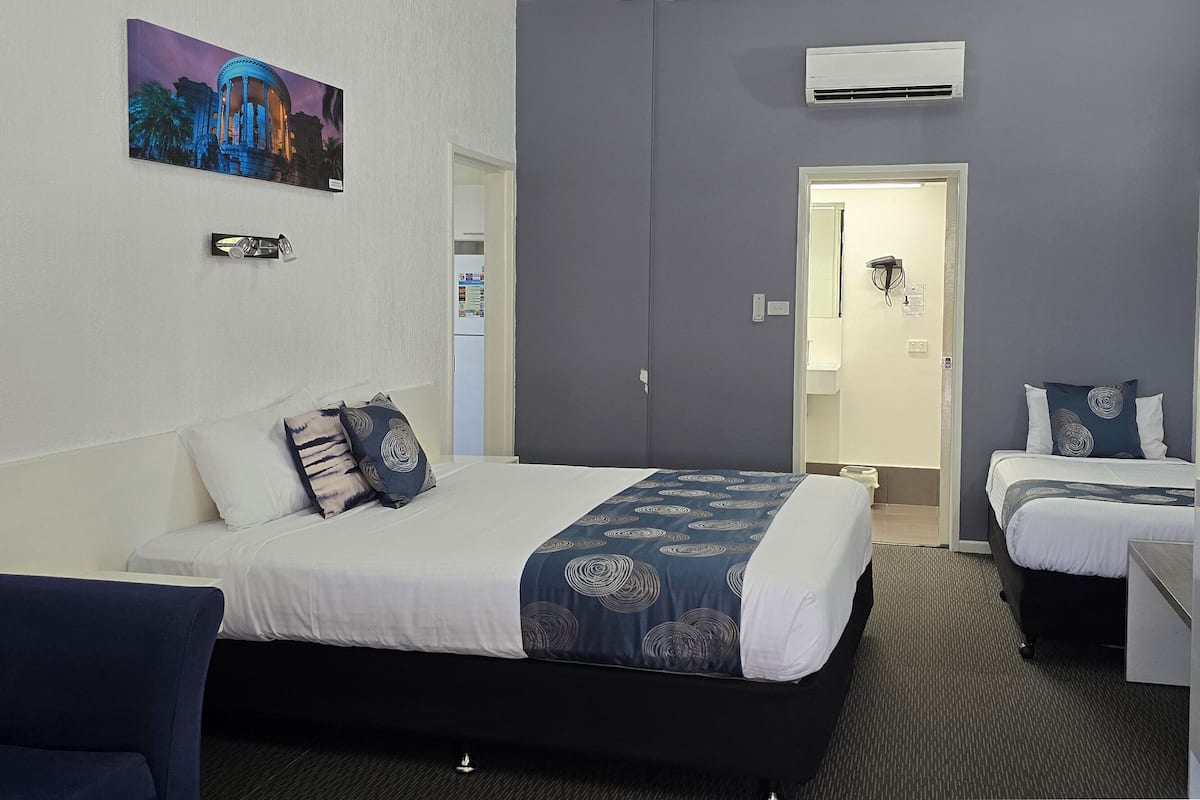 A hotel room with a large bed, a smaller bed, a desk, and a chair. There is a painting on the wall and an air conditioning unit above the door.