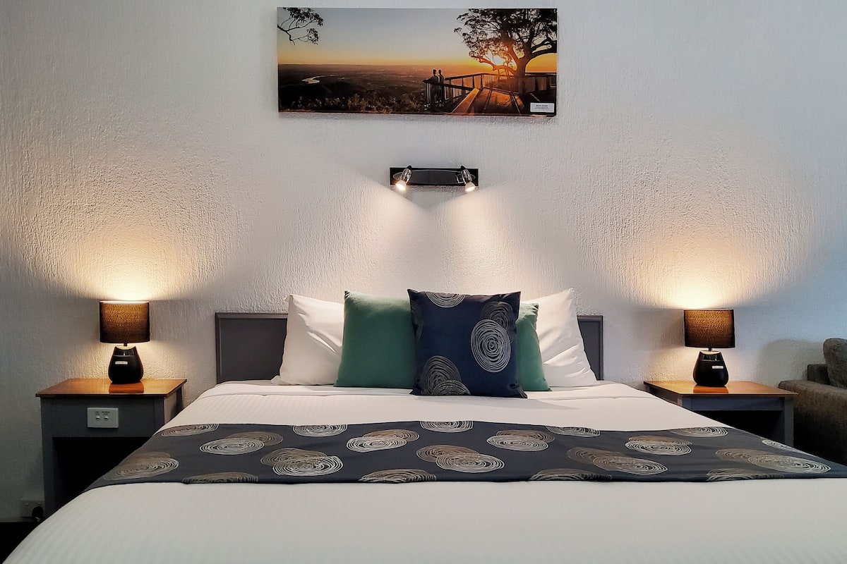 A neatly made bed with a patterned bedspread, two bedside lamps, and a painting of a sunset on the wall.