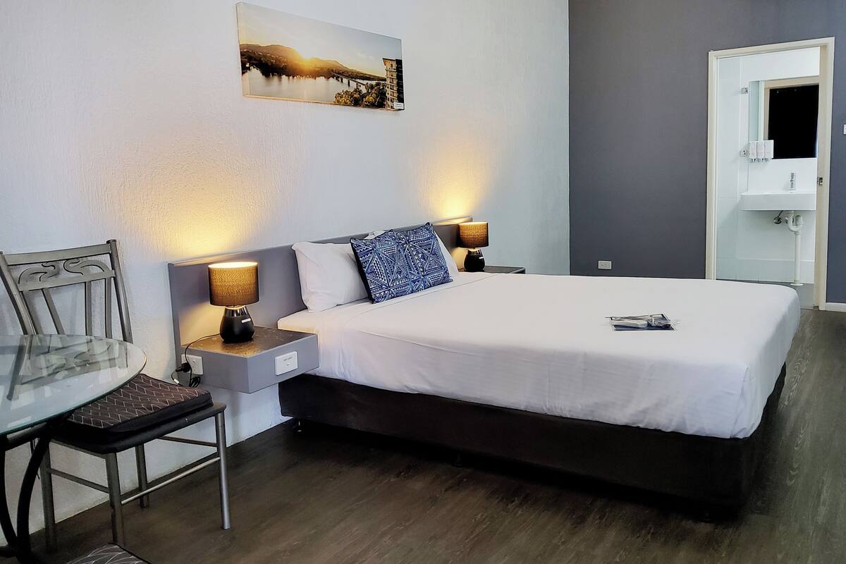 A neatly arranged hotel room with a bed, bedside lamps, a chair, a small table, and a painting on the wall.