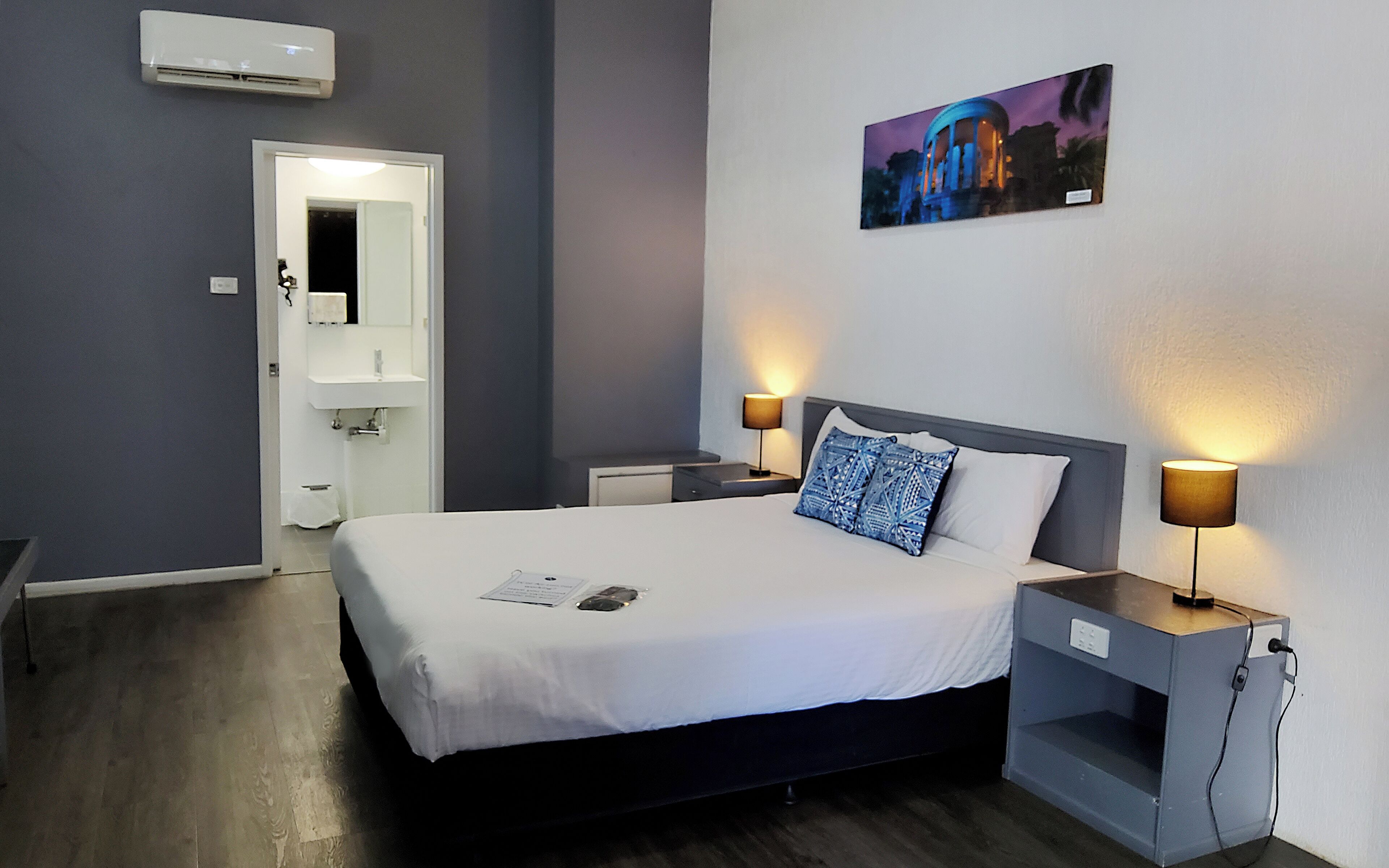 A hotel room with a bed, bedside lamps, a nightstand, a wall-mounted air conditioner, a painting, and a door leading to the bathroom.