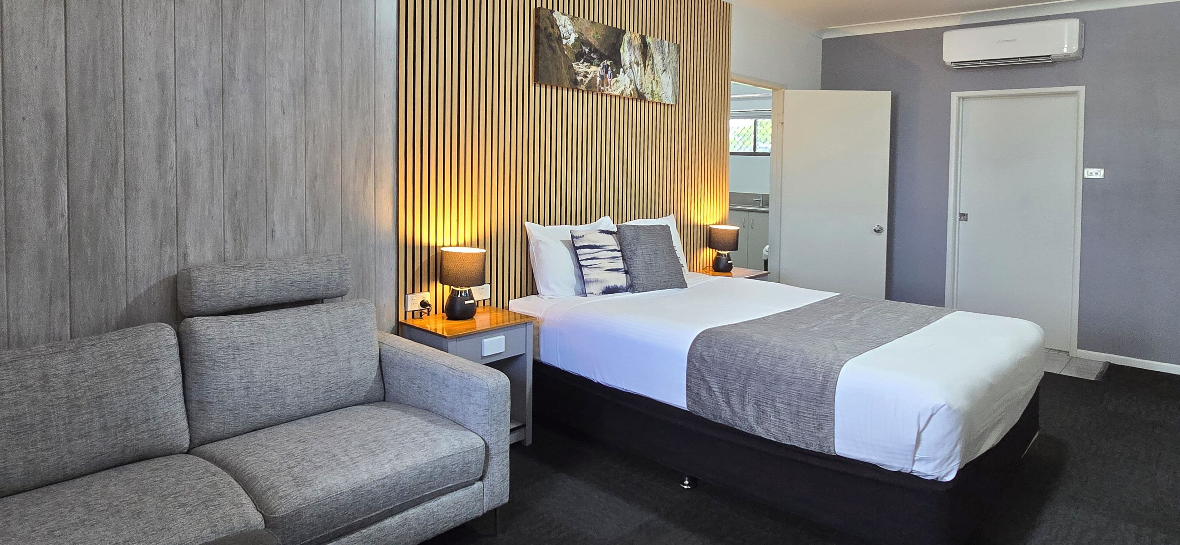 A hotel room with a bed, a sofa, two bedside tables with lamps, a wall-mounted air conditioner, and a door.