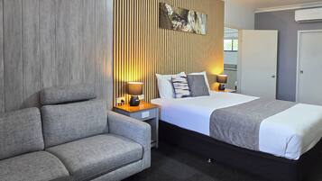 A hotel room with a bed, a sofa, two bedside tables with lamps, a wall-mounted air conditioner, and a door.