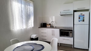 A compact kitchen with white cabinets, a microwave, a sink, and a refrigerator. There is a window with patterned curtains and a table with a placemat. - Archer Park Motel (Rockhampton)