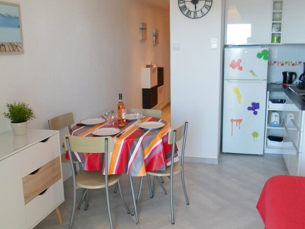 A dining area with a table set for four, a clock on the wall, and a refrigerator with colorful stickers.