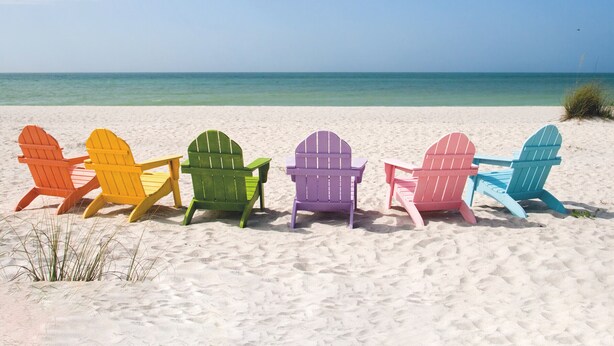 Una hilera de sillas Adirondack de colores sobre una playa arenosa, con vista despejada al océano.