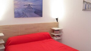 Un dormitorio con cabecera de madera, colcha roja, mesita de noche con libros y una lámpara de pared.