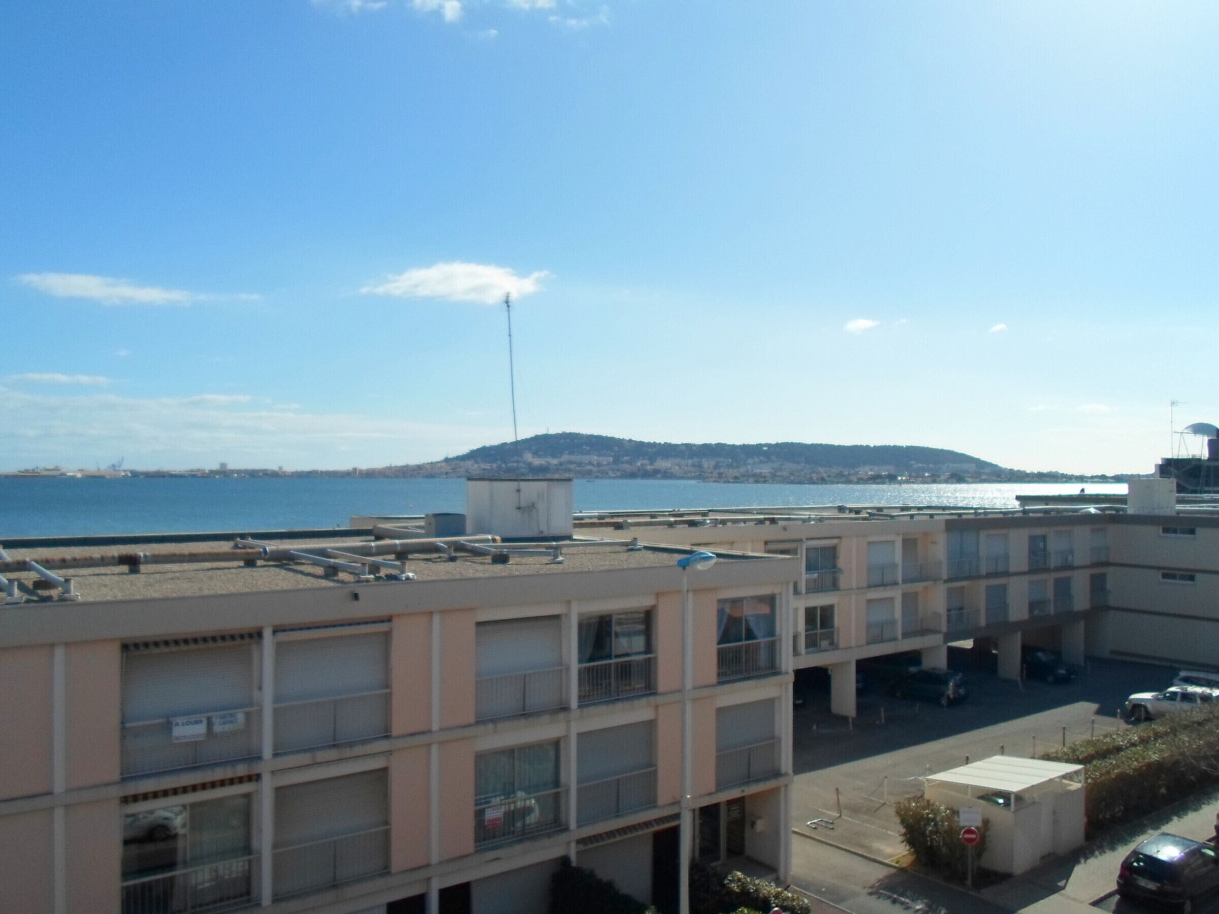Un immeuble à plusieurs étages, avec un espace de stationnement à l’avant, qui offre une vue sur une étendue d’eau et une colline lointaine.