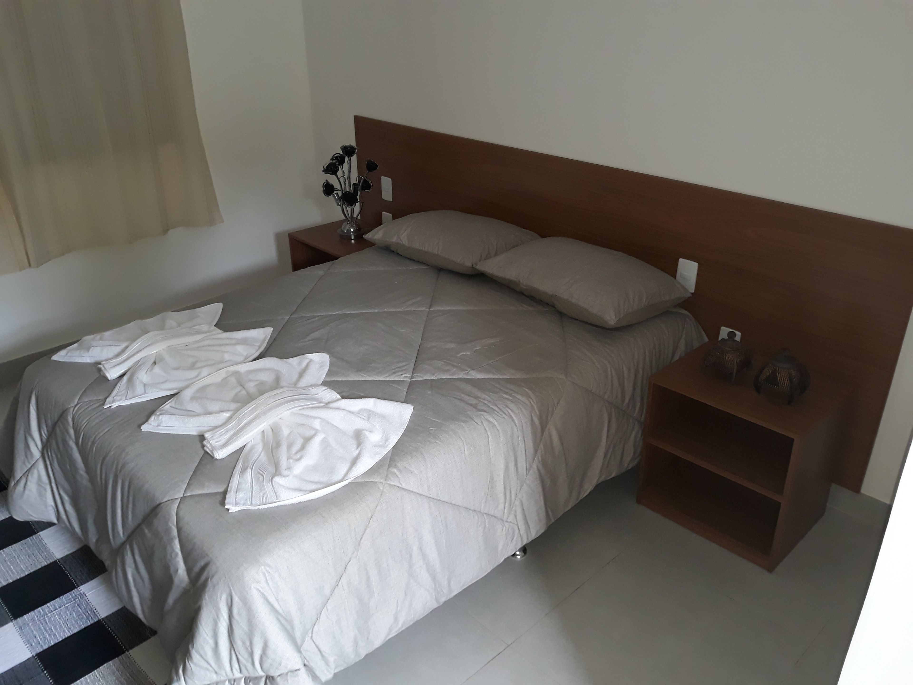 A neatly made bed with a beige duvet, pillows, and folded white towels on the bed. There is a nightstand with a vase of dried flowers and a small decorative item.