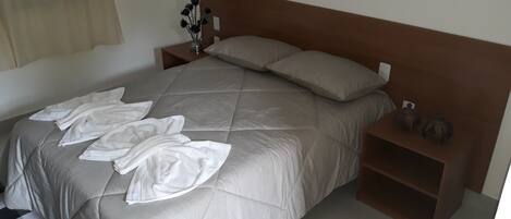 A neatly made bed with a beige duvet, pillows, and folded white towels on the bed. There is a nightstand with a vase of dried flowers and a small decorative item.