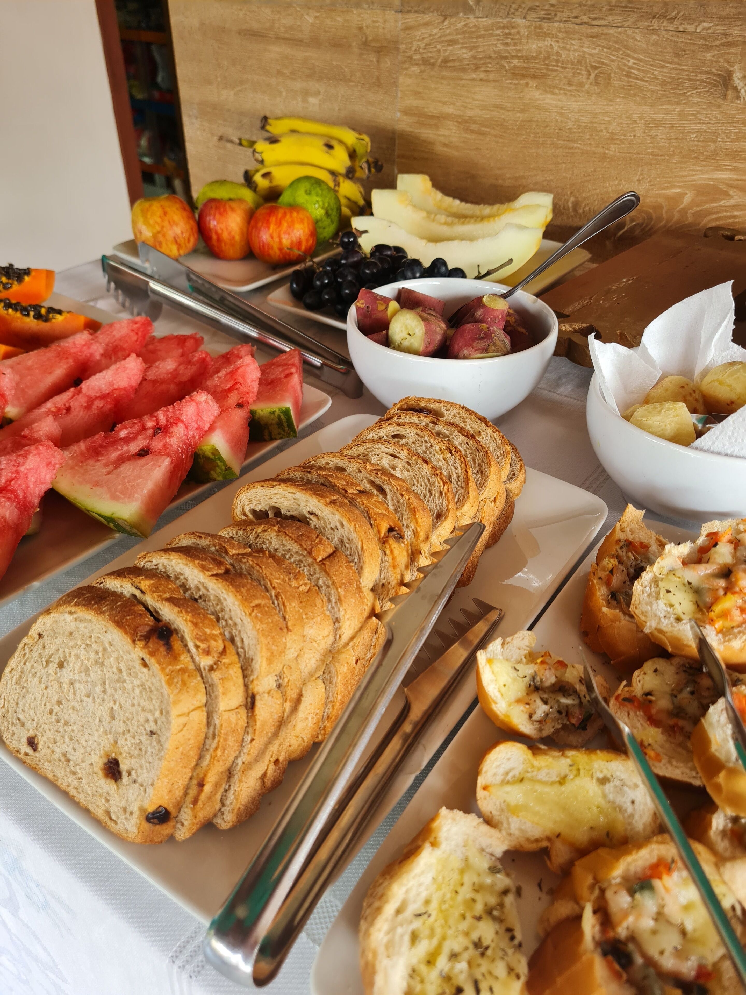 Un bufé de desayuno con pan en rodajas, sandía, uvas y diversas frutas.