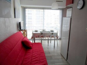 Ein kompakter Wohnraum mit einem roten Sofa, einem Esstisch mit Stühlen, einem Fernseher und einem Kühlschrank.