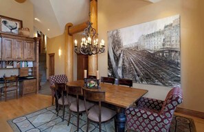 A dining area with a wooden table, chairs, a chandelier, and a large painting of a bridge.