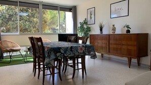 Ein Essbereich mit einem Tisch für vier Personen, umgeben von Holzsesseln. Ein Holzschrank mit Dekorationen steht daneben, und ein großes Fenster bietet Blick ins Grüne.