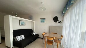 A compact living space with a black sofa, a small dining table with chairs, a ceiling fan, and a mounted TV.