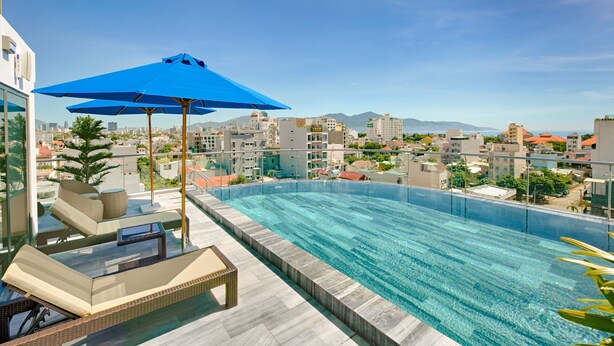 A rooftop infinity pool with lounge chairs and a blue umbrella, overlooking a cityscape with buildings and mountains.