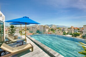Ein Rooftop-Infinity-Pool mit Liegestühlen und einem blauen Sonnenschirm mit Blick auf eine Skyline aus Gebäuden und Bergen.