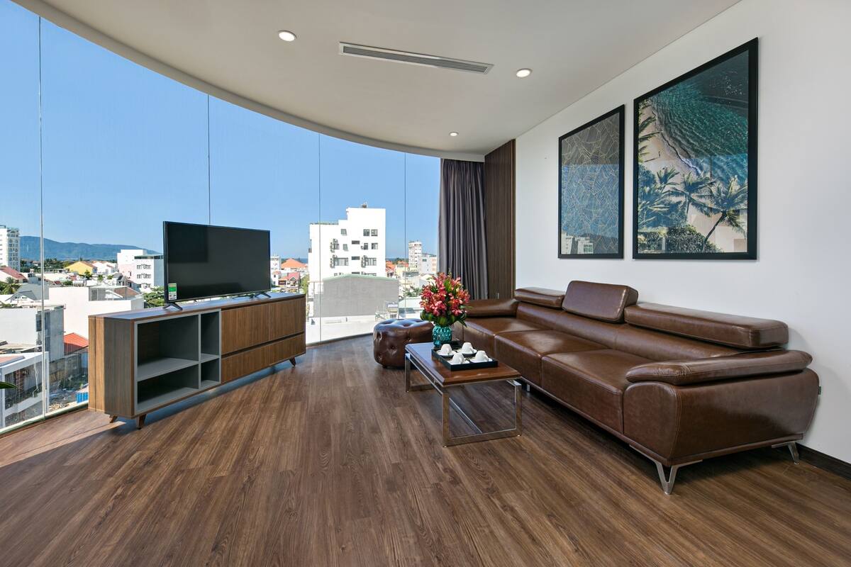 Ein modernes Wohnzimmer mit einem großen Flachbildfernseher, einem braunen Ledersofa, einem Holztisch und Blick auf die Stadt durch große Fenster.