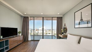 A modern hotel room with a large bed, a flat-screen TV, a wooden dresser, and a balcony with a city view.