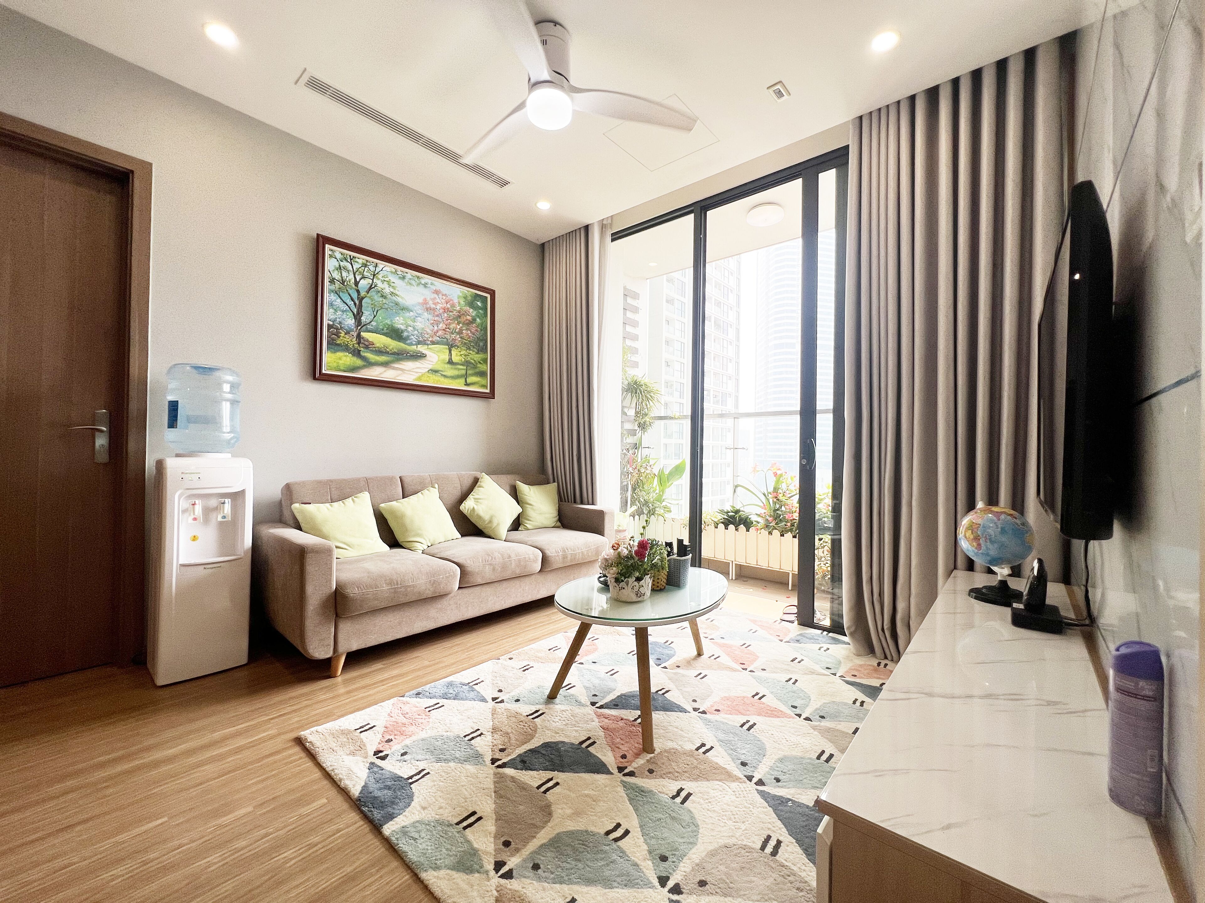 A modern living room with a sofa, a coffee table, a water dispenser, a painting, and a view through large windows.
