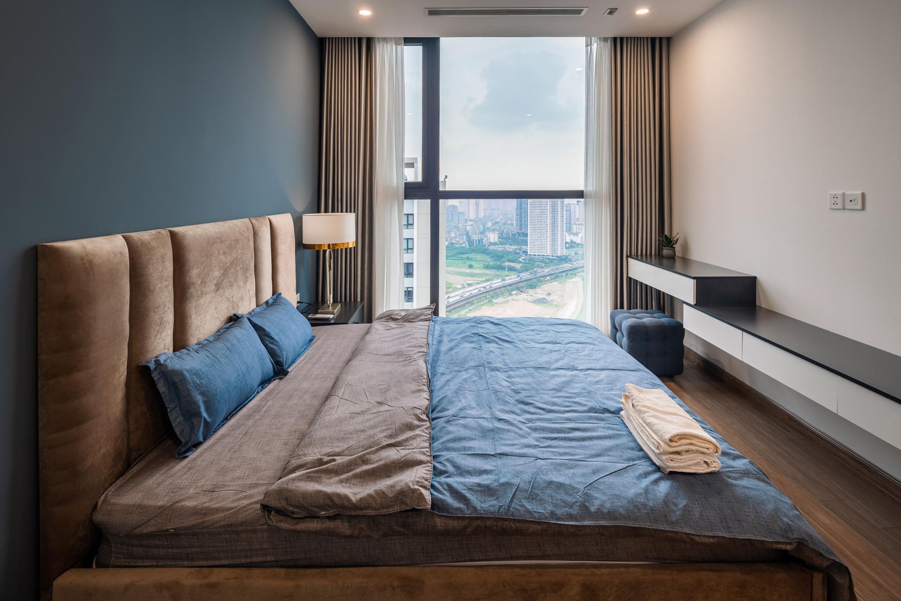 Een moderne hotelkamer met een groot bed, een nachtkastje met een lamp, uitzicht op de stad door grote ramen en een kleine plant op het nachtkastje.