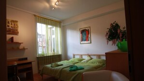 Een slaapkamer met een bed, een bureau, een stoel, een raam met gordijnen, een schilderij aan de muur en een plant.