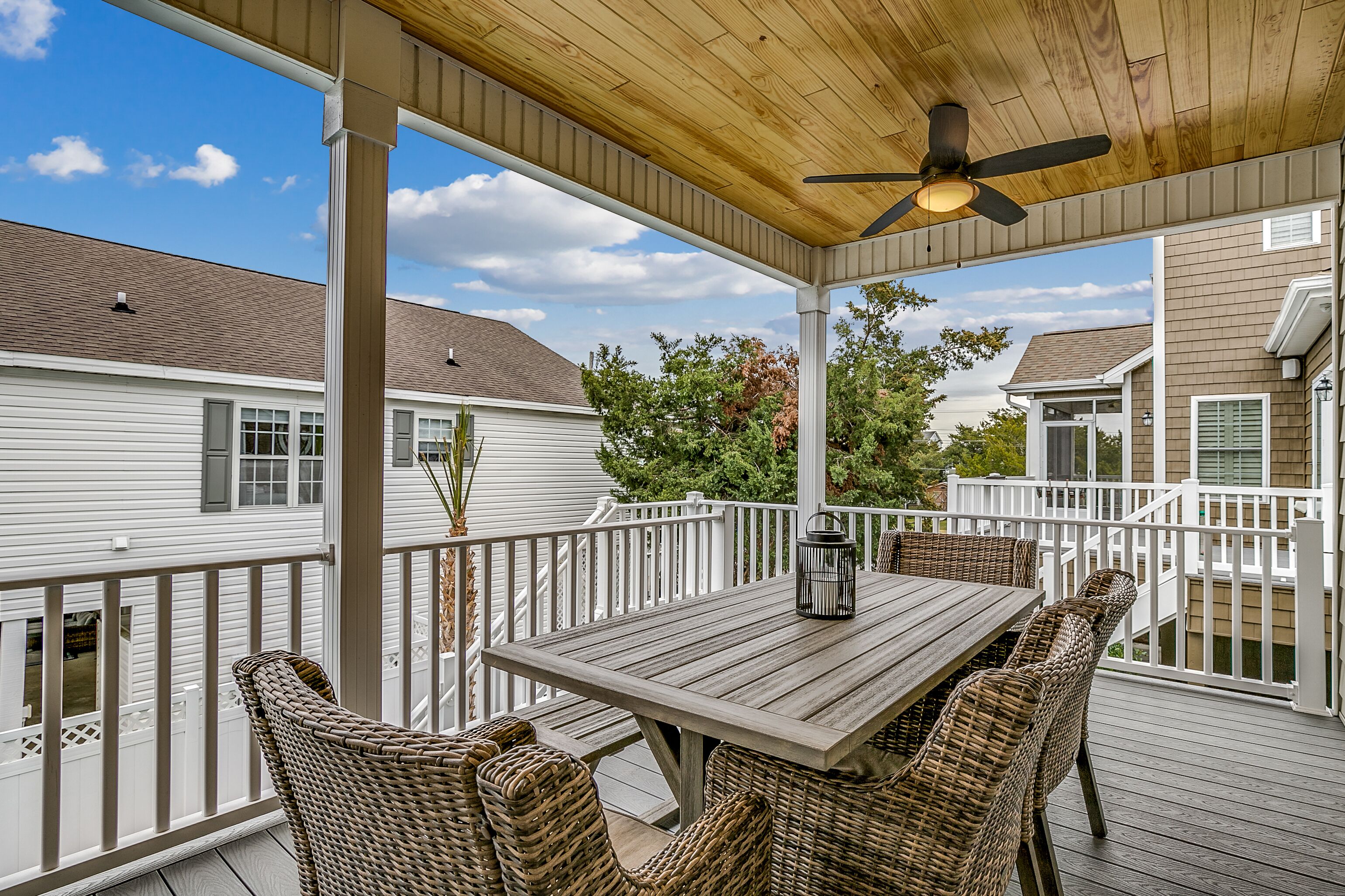 A covered outdoor deck with a wooden table and wicker chairs, surrounded by white railings and a view of neighboring houses.