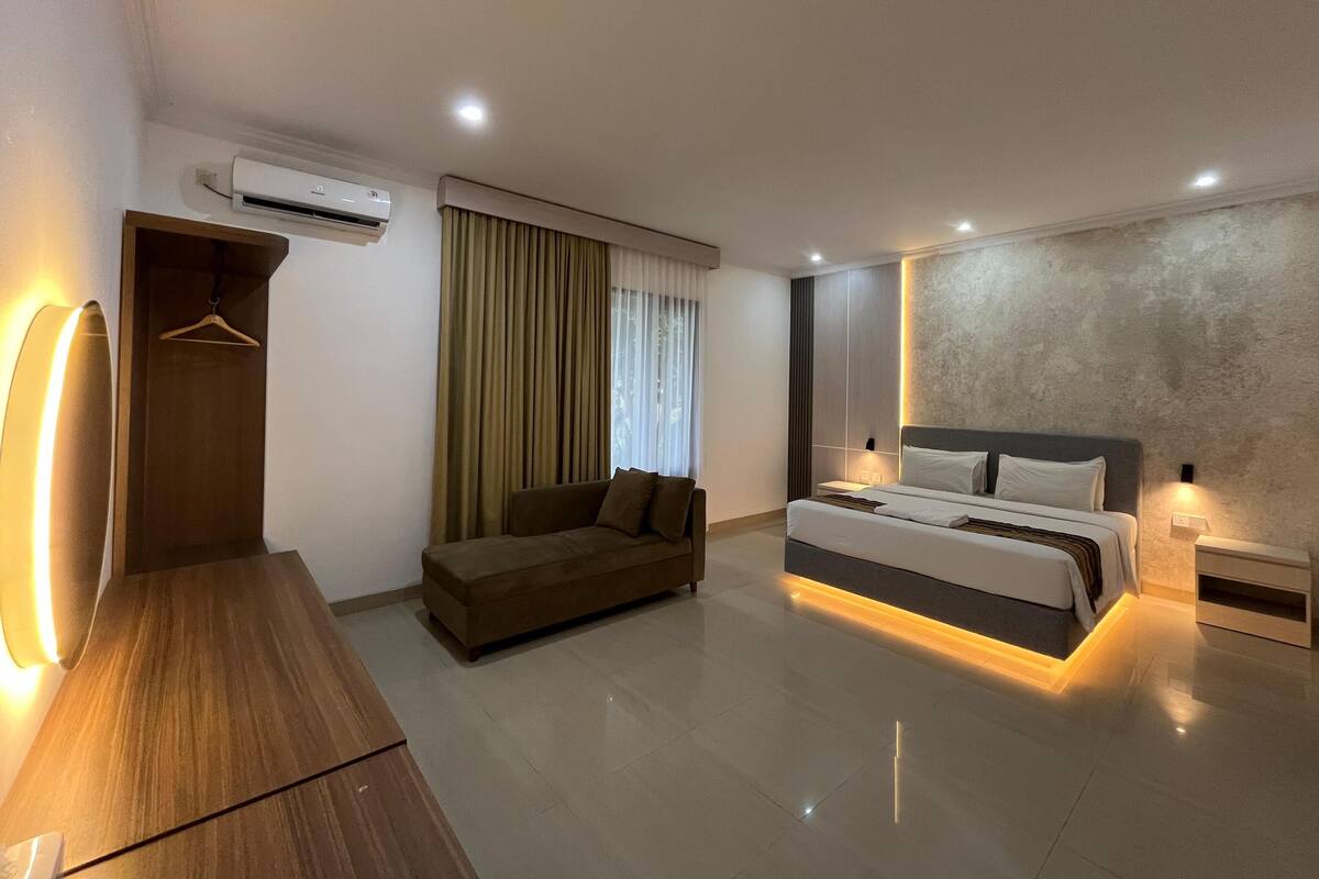 A modern hotel room with a bed, a sofa, a wooden desk, and an air conditioning unit.