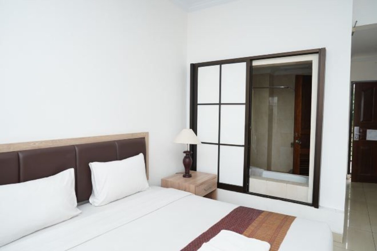 Habitación de hotel con cama, mesita de noche con lámpara y puerta corredera hacia el baño.