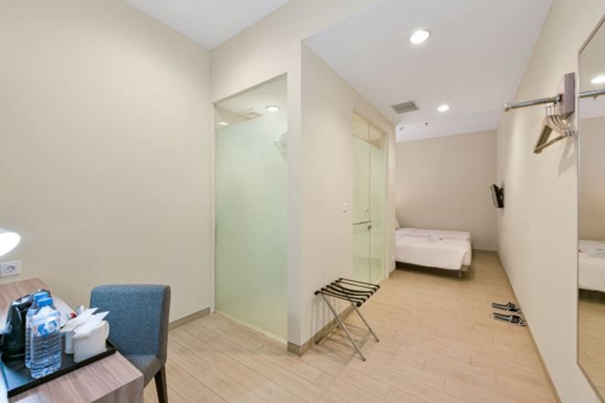 A hotel room with a bed, a small table with a lamp, a chair, a towel rack, and a glass partition.
